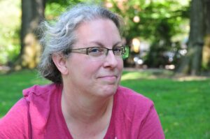 A close-up photo of Kymm with her hair pulled back. She is wearing a pink t-shirt and glasses, and has a smirk on her face.