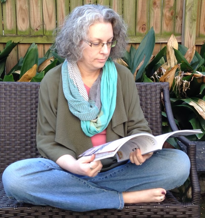 Closeup of Kymm sitting on a bench in a garden reading a book barefoot, in a square frame